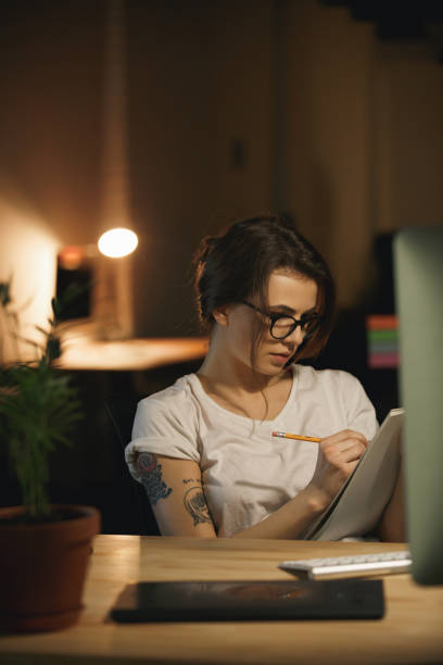 istock Concentrated young lady designer sitting indoors at night writing notes. 1356227498