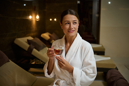Attractive elegant Caucasian young woman in white bathrobe, holding a glass cup of hot healthy herbal drink, looking aside, sitting on chaise lounge by hot tub, enjoying spa procedures in spa salon