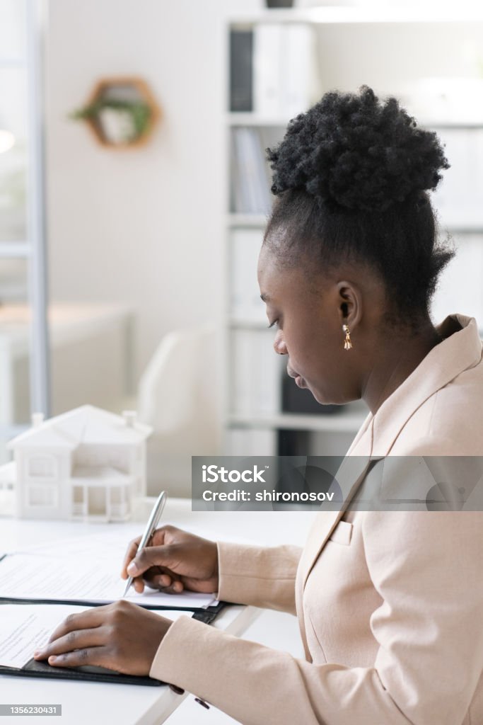Blackwoman Signing Mortgage Document Serious young Blackwoman with curly hair sitting at table in bank and signing mortgage document 20-24 Years Stock Photo