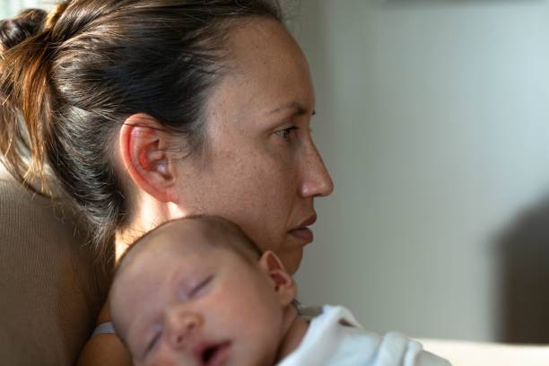 A sad mother sitting at home suffering from postpartum depression. Motherhood problems. A unhappy mom holding her baby at home sitting on the couch thinking. postpartum depression stock pictures, royalty-free photos & images