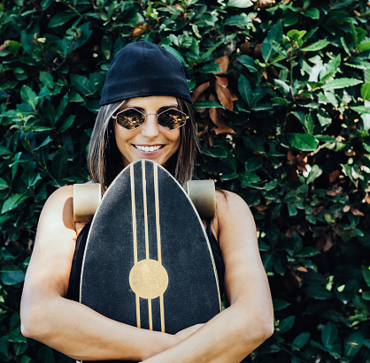 a girl with sunglasses and a black cap looks at the camera and smiling holding a skateboard in her arms. concept of happiness and free time.