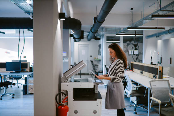 Woman using the photocopier machine at the office Portrait of a mid age woman in her 40s, finishing work. She is back into the office after the coronavirus pandemic. computer printer stock pictures, royalty-free photos & images