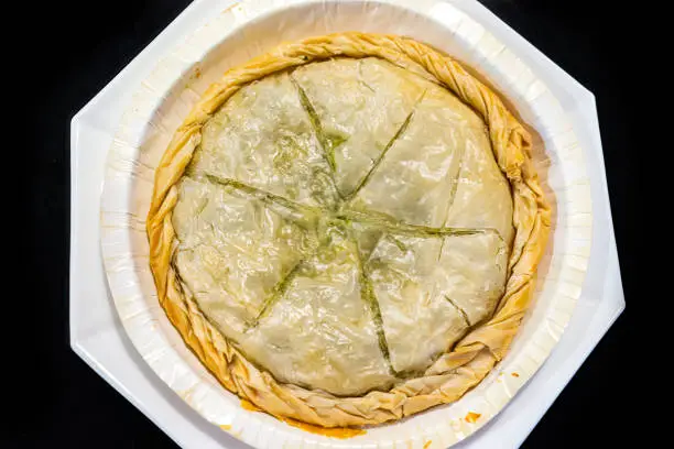 Flat top lay view looking down closeup of filo dough Spanakopita whole pie on white plate isolated black background as traditional greek food made with spinach and cheese baked