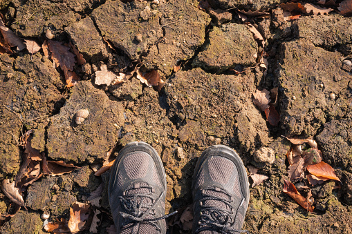 Man's feet in hiking boots on cracked soil on the lake shore due to drought. Background for drought, global warming and environmental problems. Top view.