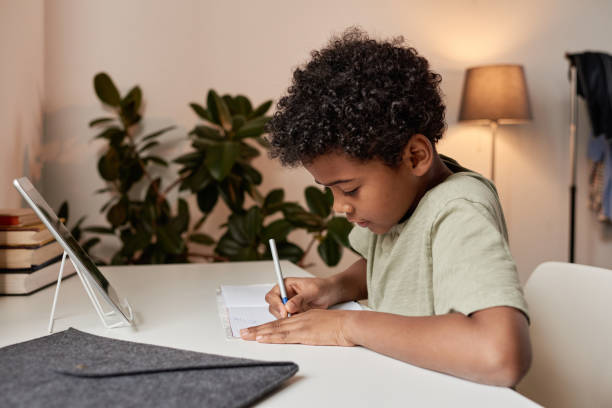 Serious curly-haired black boy sitting at desk and making notes in workbook during online lesson at home