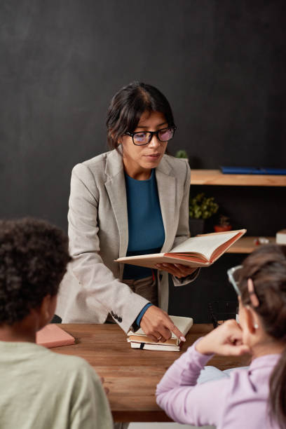 giovane insegnante serio di razza mista che tiene un libro di testo aperto e spiega un nuovo argomento ai bambini in classe - open book teaching table foto e immagini stock