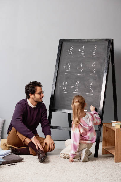 istock Girl with ponytail pointing at blackboard while learning to add and subtract numbers with tutor 1356248155