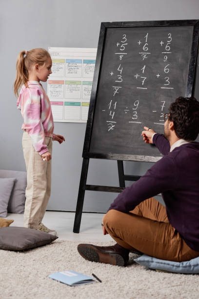 istock Blond-haired girl in hoodie standing in front of blackboard and solving math problem with tutor 1356249184
