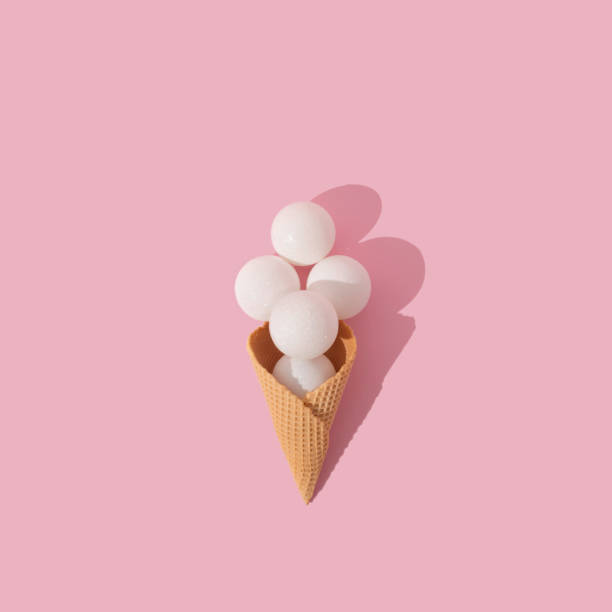 istock Ice cream cone with white  christmas baubles under direct light on pastel pink background. Christmas and new year celebration concept, flat lay perspective, square ratio. 1356253522