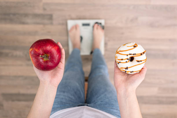 una mujer se para en la balanza, sostiene una rosquilla y una manzana en sus manos, vista superior. - weight scale apple comparison balance fotografías e imágenes de stock
