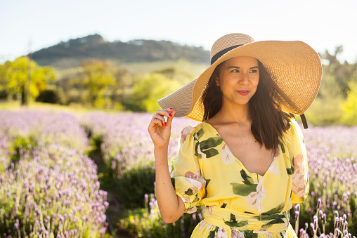 Young woman wearing a sun hat smiling while standing in a field of lavender on a sunny afternoon in summer