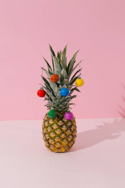 istock Pineapple decorated as christmas tree with christmas baubles colored as rainbow flag on white and pastel pink background. LGBT christmas celebration concept. Close up perspective. 1356253706