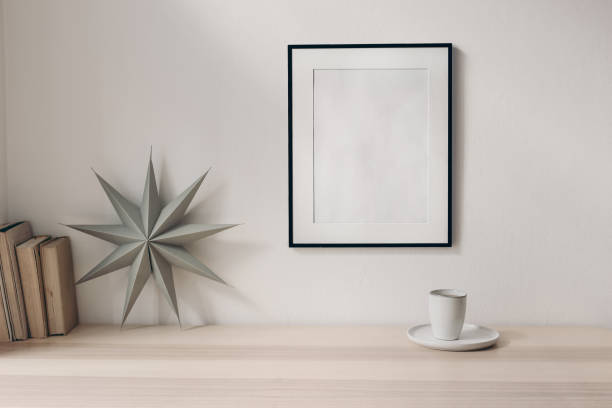 Empty black picture frame on white living room wall. Stationery mockup scene with grey folded paper star, cup of coffee and books on wooden table backgroum. Christmas decoration, winter composition. stock photo