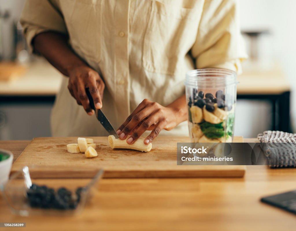 Close Up of Woman Hands Cutting Banana on a Cutting Board Unrecognizable African American woman making healthy breakfast on a kitchen table. Healthy Eating Stock Photo