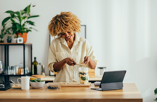 Afro-American woman with a big smile on her face is cutting fruit to prepare a healthy breakfast while listening to a video on a digital tablet. She has a digital tablet, bowl with fruit, cutting board and a clot on the table in front of her.