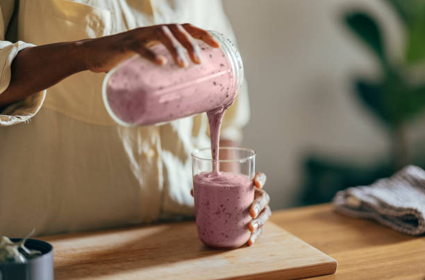 Anonymous Afro-American Woman Pouring a Smoothie Into a Glass Close-up of hands of an Afro-American woman wearing beige shirt and pouring smoothie into a glass on a kitchen table. There is a part of a blender, a cutting board and a cloth on the desk in front of her. In the corner of a room sits a big plant. smoothie stock pictures, royalty-free photos & images