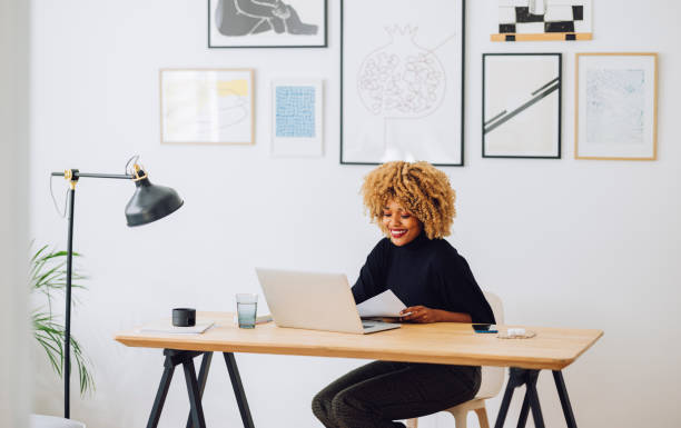 femme afro-américaine souriante assise au bureau dans son bureau et regardant un ordinateur portable - grand angle photos et images de collection