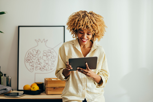 Happy Afro-American woman is standing in a room and leaning against a drawer while looking at her digital tablet. She has jeans and a beige shirt on. There is fruit on a drawer behind her.