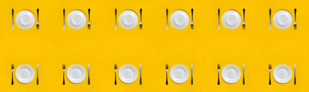 A set of twelve empty white plates, forks and knives lie on a yellow tablecloth. Minimalistic table setting, top view. The last supper concept. Copy space. stock photo