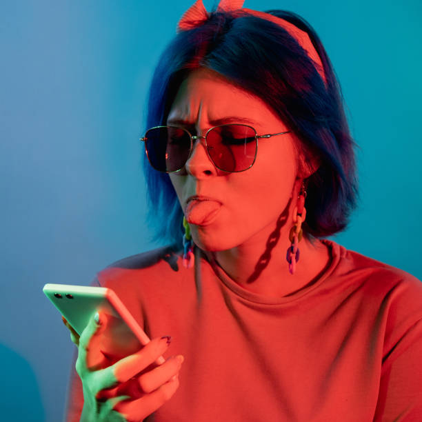 haters harassment online spam woman phone neon Haters harassment. Online spam. Low internet. Annoyed woman sticking tongue out at phone in red neon light isolated on blue background. phone spam photos stock pictures, royalty-free photos & images
