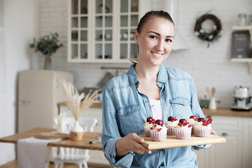 Work in confectionery workshop. Handmade cupcakes. Smiling pretty young woman pastry chef, demonstrating tasty fresh cupcales with berries to camera