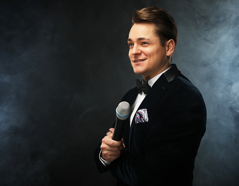Stylish young man in a tuxedo holding a microphone, posing against a dark background with smoke, actor, singer, show, host of the event.