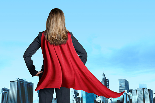 A rear view of a businesswoman wearing a red cape as she looks out toward the skyline of a large city.
