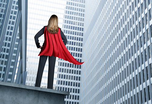 A rear view of a  businesswoman wearing a red cape standing with her hands on her hips in front of the glass and steel of big city skyscrapers.