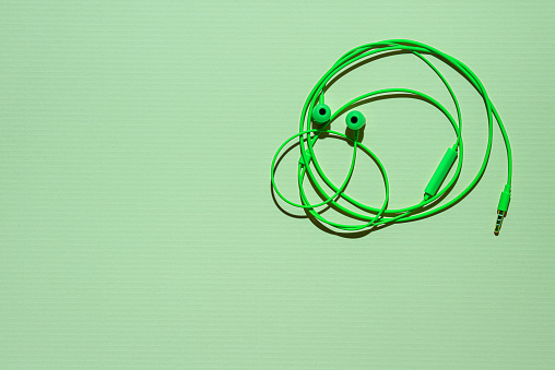 Bright green earphones with microphone and 3,5 mm mini-jack connector on green textured paper background. Wire in shape of funny round face of human with eyes and nose. Large copy space. Flat lay.