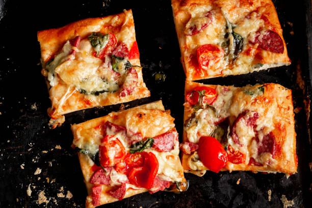 スライスした正方形のピザ、パイ、トマト、ソーセージ、チーズをオーブンで調理した黒いベーキングシート。 - tomato sauce domestic kitchen meat cheese ストックフォトと画像
