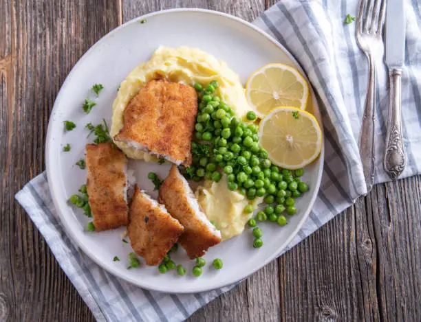 Traditional fresh cooked and homemade breaded fried fish with mashed potatoes and buttered green peas. Served on a plate isolated on wooden table. Closeup and overhead view