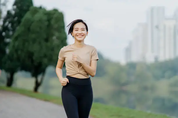 Beautiful young healthy Asian woman running in the park. Happy female runner jogging in the morning in park.