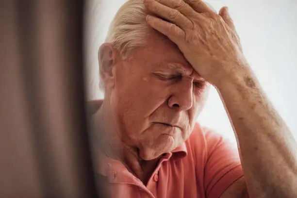 Close up of a senior man who has a chronic illness/Alzheimer's disease at his home in the North East of England. He is holding his forehead with a negative expression, experiencing anxiety/pain.