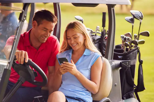 Photo of Couple Sitting In Golf Buggy On Course With Woman Using Mobile Phone