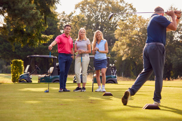 mature and mid adult couples standing by golf buggy watching man hit tee shot - golf four people young adult playing imagens e fotografias de stock