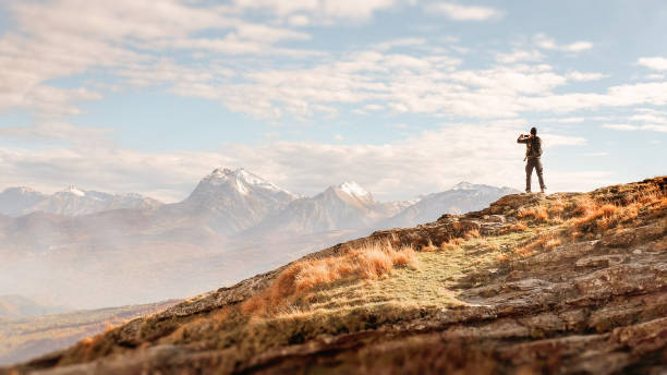 Male traveler on top of a rock taking pictures of majestic mountain landscape Adult male hiker standing on top of a rock taking pictures of majestic mountain landscape with a smartphone. Warm light with beautiful sky. Total freedom and exploration. photo messaging stock pictures, royalty-free photos & images