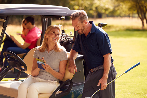 Mature And Mid Adult Couples In Buggies Playing Round On Golf Together