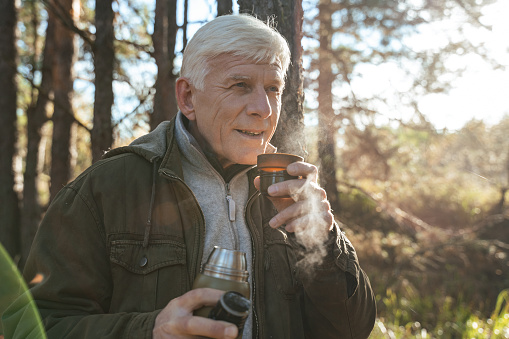 Grey haired man looking away and holding cup with hot tea while enjoying of the aromatic beverage and feeling calm at the morning autumn forest