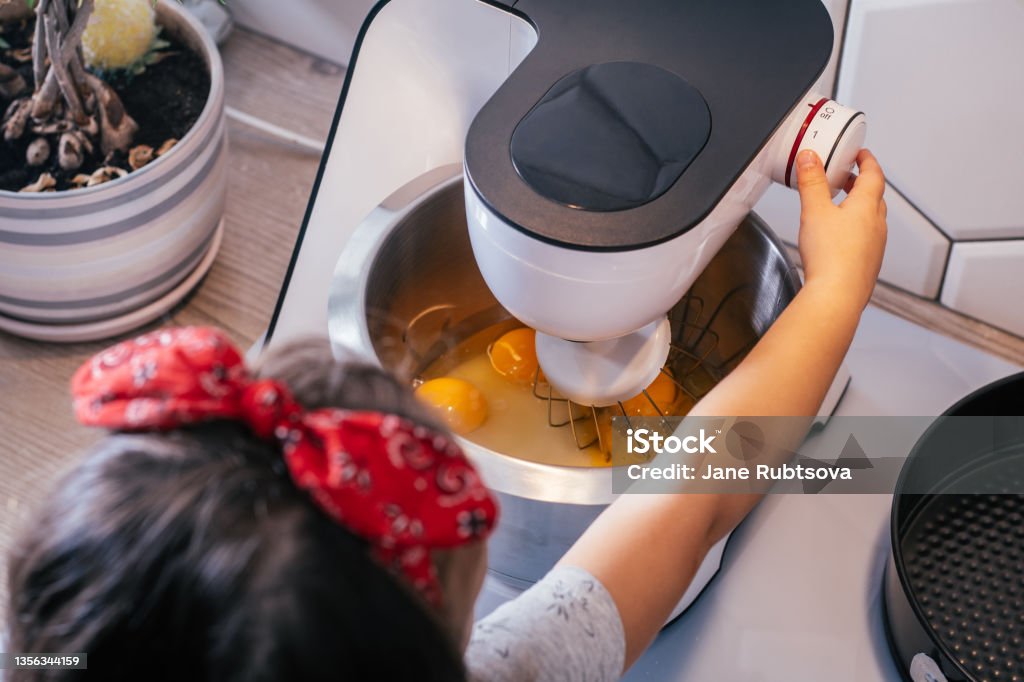 Little dark-haired girl 3 years old in red headband bakes apple pie in kitchen. Child turns on planetary mixer. Top view Little dark-haired girl 3 years old in red headband bakes apple pie in kitchen. Child turns on planetary mixer. Children help on household chores. Kid cooking food. Top view Baker - Occupation Stock Photo