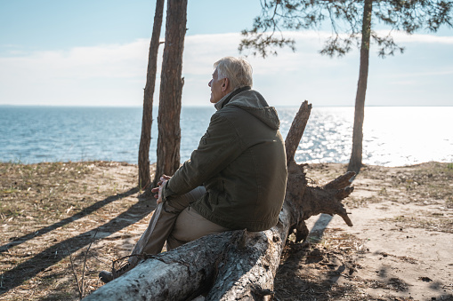 Great moment. Side view of the calm senior man sitting at the tree log at the seashore and looking at the water surface with pleasure emotions