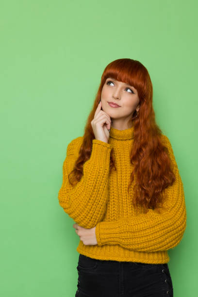 Cute red haired young woman in yellow turtleneck sweater is looking up and thinking. stock photo