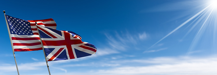 Flags of the United States of America and United Kingdom, with flagpole, blowing in the wind on a blue sky with clouds and copy space and sunbeams. Photography.