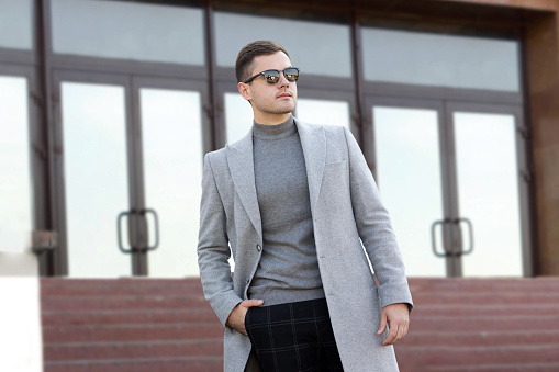 Handsome young man in gray autumn coat stands on the steps of a building