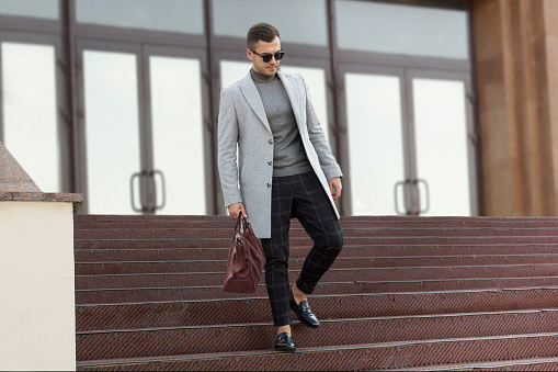 Handsome young man in gray autumn coat with leather bag goes down the steps of the building