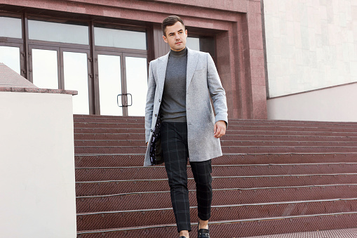 Handsome young man in gray autumn coat goes down the steps of the building