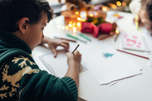 Boy drawing a Christmas wish list, sitting in a living room in front of the Christmas tree