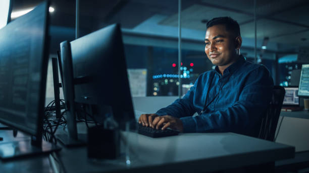 istock Night Office: Portrait of Handsome Indian Man in Working on Desktop Computer. Digital Entrepreneur Typing, Creating Software, e-Commerce App Design, Programming. Thoughtful Happy Man Finding Solution 1356364201