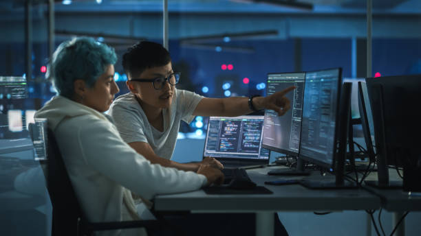 istock Teamwork In Diverse Office at Night: Project Manager Talks with Creative Solutions Specialist, They Discuss App Code Computer. Stylish Professionals Programming, Create e-Commerce Software 1356364226