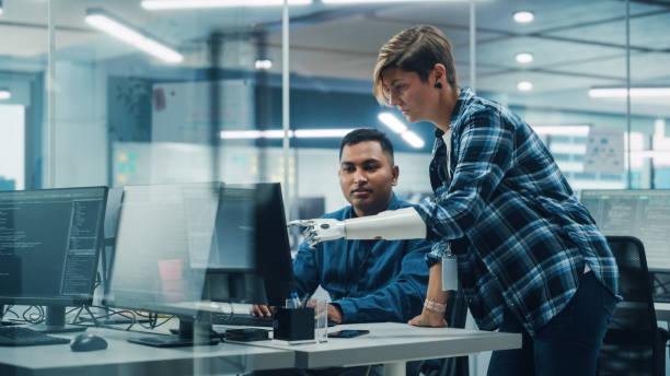 trabajo en equipo en una oficina inclusiva diversa: gerente de proyecto de mujer con discapacidad con brazo protésico habla con especialista indio que trabaja en computadora de escritorio. los ingenieros de software profesionales crean una aplicación - special needs fotografías e imágenes de stock