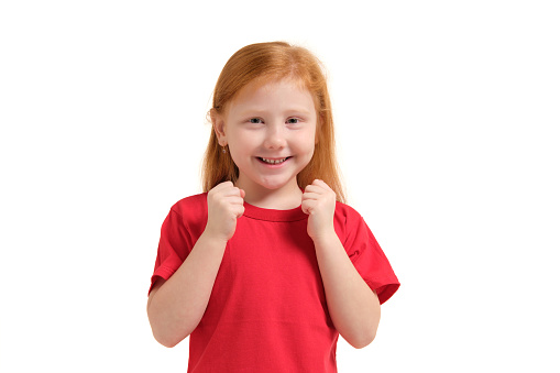 Cheerful little girl, look so excited she is raising her fists up, isolated on white background. Emotions of a redhead girl in a red t-shirt